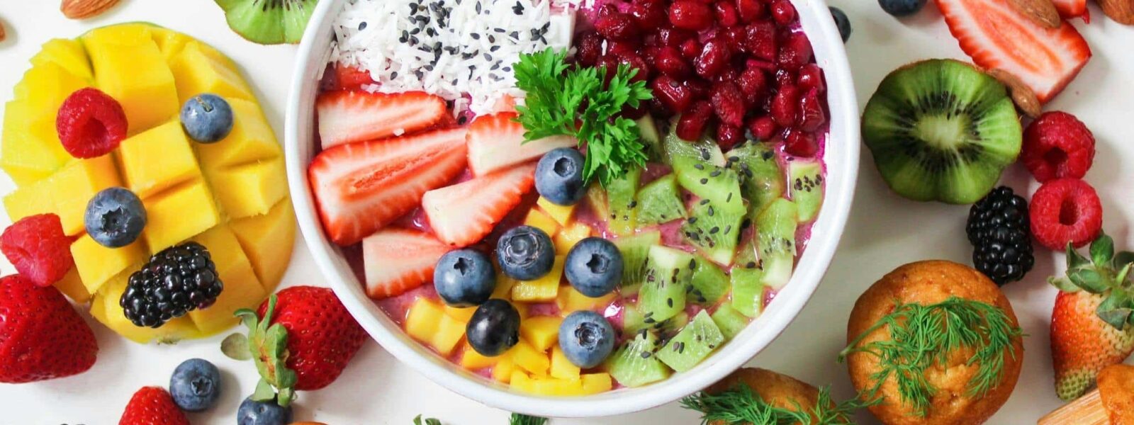 Antiinflammatorisk Smoothie Bowl – Sunt Kosthold ved Lipødem Fargerik smoothie bowl med friske bær, kiwi, mango og kokos omgitt av tropisk frukt, nøtter og muffins på hvit bakgrunn
