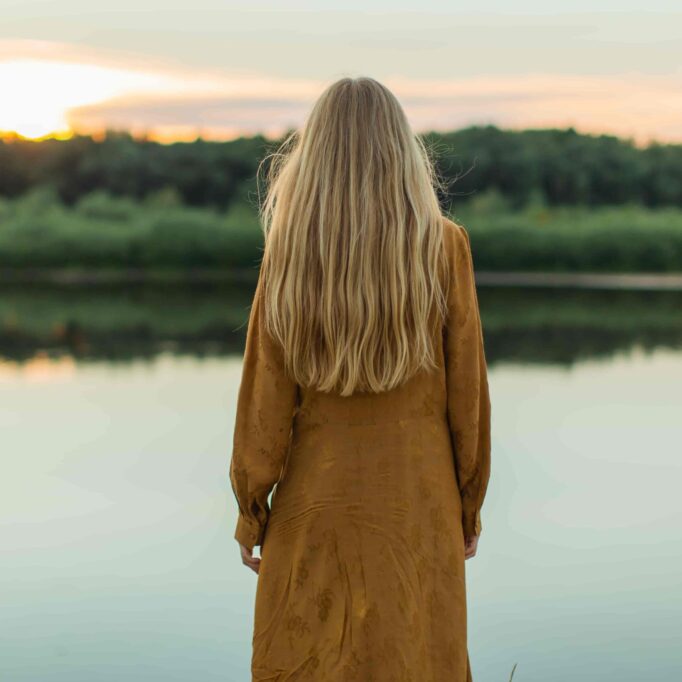 En dame med langt hår i brun frakk står med ryggen til og ser utover et vann.